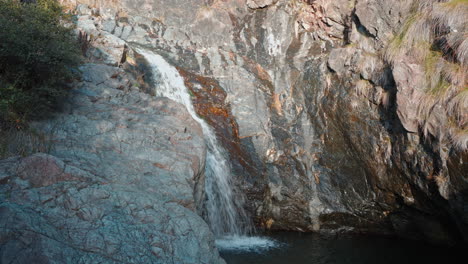 Small-waterfall-cascading-down-a-rocky-cliff-into-a-tranquil-pool,-sunlight-enhancing-the-serene-landscape