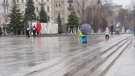 Children-walk-across-Independence-monument-wrapped-in-blue-yellow-flag-with-umbrella-on-rainy-day