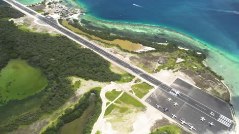 Los-roques'-landing-strip-by-clear-turquoise-waters-and-lush-greenery,-bright-daylight,-aerial-view