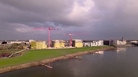 Aerial-approach-over-river-IJssel-towards-Dutch-new-build-neighbourhood-Noorderhaven-with-red-cranes-rising-above-construction-site