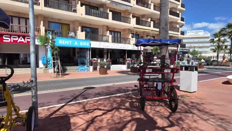Cala-Bona-rental-bikes,-pedals-on-a-sunny-promenade-with-hotels,-shops,-palm-trees,-Majorca,-Spain
