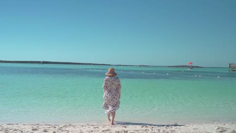 Woman-wearing-a-hat-and-a-dress-walks-into-beautiful-sea-slow-backshot