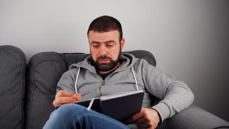 Man-Writing-On-Notebook-While-Sitting-On-The-Sofa