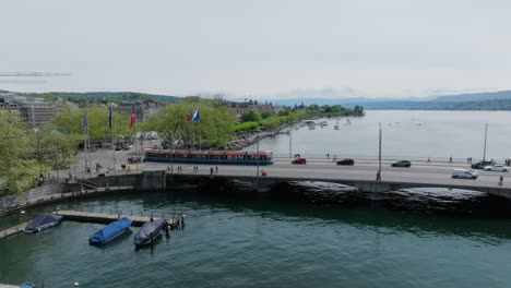 Slow-forward-moving,-panning-drone-shot-showing-the-main-bridge-of-the-city,-Swiss-and-EU-flags,-pedestrians,-transport-and-Lake-Zurich