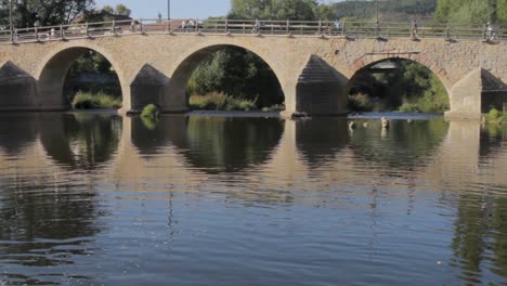 Old-stone-bridge-reflected-in-tranquil-river-water,-sunny-day,-green-foliage-in-background,-pedestrians-visible