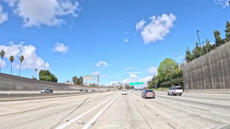 Sunny-day,-driving-on-Los-Angeles-freeway,-palm-trees-lining-the-road,-fluffy-clouds-in-blue-sky,-time-lapse