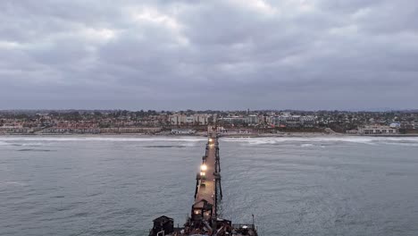 Oceanside-California-Pier-Fire-Damaged-Restaurant-Drone-Vertical-Drop-In-Reveal-Calm-Ocean-Cloudy-Sunrise-With-Shore-in-Background