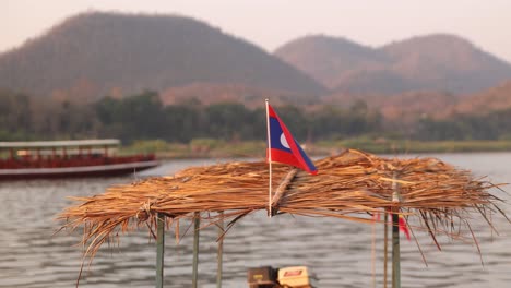 Bandera-De-Laos-Ondeando-Al-Viento-Sobre-El-Techo-De-Paja-Del-Barco-En-Luang-Prabang,-Laos-Viajando-Por-El-Sudeste-Asiático