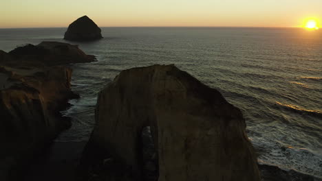 Haystack-rock-in-distance-past-sandstone-eroded-arch-of-Cape-Kiwanda-at-sunset,-aerial