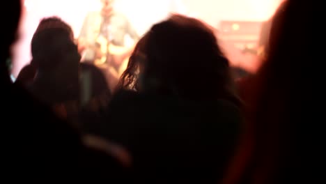 Rear-shot-of-cheering-people-celebrating-and-jumping-indoors-in-front-of-stage,-blurred-handheld-shot