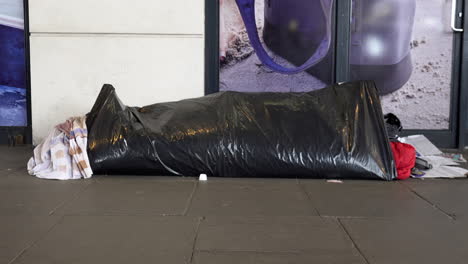 A-person-walks-past-a-homeless-person-asleep-on-a-pavement-in-a-sleeping-bag-that-is-wrapped-up-in-black-plastic-sheeting