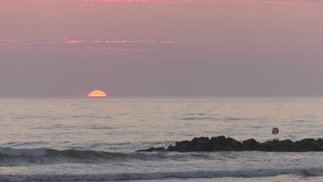 Die-Sonne-Geht-über-Dem-Meereshorizont-In-Den-Blassen-Morgenhimmel-Auf,-Wellen-Brechen-An-Den-Felsen