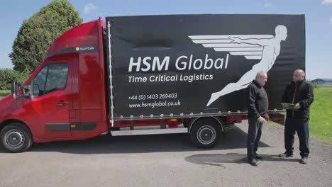 Two-men-discuss-logistical-details-next-to-HSM-Global-delivery-truck,-pan-shot