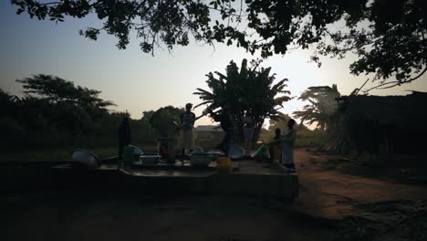 Afrikanische-Frau-Pumpt-Wasser-Aus-Einem-Wohltätigkeitsbrunnen,-Der-Bei-Sonnenuntergang-In-Afrika-Von-Menschen-Umgeben-Ist---Weitwinkelaufnahme---Zeitlupe