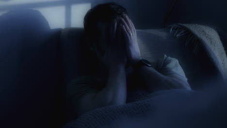 Man-covering-face-with-hands-on-couch,-tense-atmosphere,-blue-tones,-evening