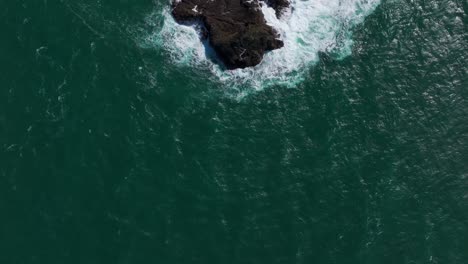Aerial-drone-view-looking-down-at-a-rock-island-as-waves-crash-over-it