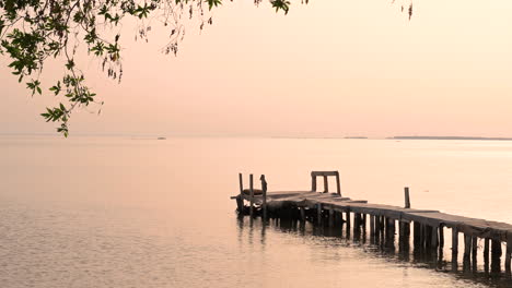 Wooden-old-pier-on-calm-water-waves-on-the-sea-with-wind-blowing-green-leaves-frame-over-small-island-at-sunset-sky,-Bahrain