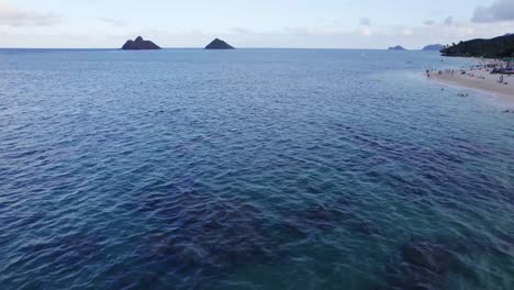 Drohnenaufnahmen-über-Dem-Pazifischen-Ozean-In-Der-Nähe-Von-Oahu,-Hawaii,-Mit-Weißem-Sandstrand-Auf-Der-Rechten-Seite-Und-Zwei-Kleinen-Inselchen-Am-Horizont-Und-Korallenriffen-Auf-Dem-Meeresboden