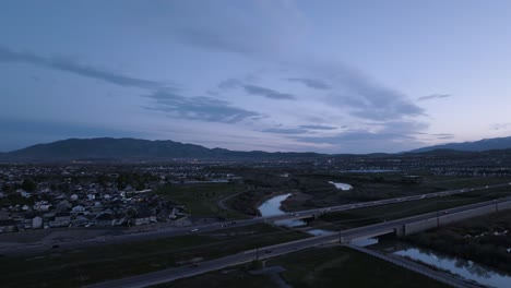 Highway-across-a-river-in-a-valley-beneath-the-mountains-at-twilight---pullback-aerial-reveal
