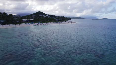 drone-footage-Lanikai-Beach-on-the-island-of-Oahu-showing-reefs-on-the-ocean-floor-with-blue-green-water-and-a-small-beach-town-lining-the-coast-and-white-sand-beach,-island-mountains-on-the-horizon