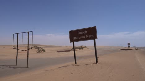 Namensschild-Für-Den-Dorob-Nationalpark-Neben-Einem-Alten-Kaputten-Schild,-Sand-Weht-An-Einem-Sonnigen-Tag-Auf-Dem-Boden