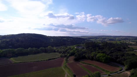 Aerial-drone-view-of-a-road-surrounded-by-fields-and-nature,-lots-of-trees-and-greenery,-a-hill-on-the-left-and-horizon,-blue-sky-with-clouds