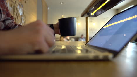 Close-up-of-a-man's-hands-writing-on-a-notebook-and-drinking-coffee,-inside-a-cozy-bar-with-people-in-the-background