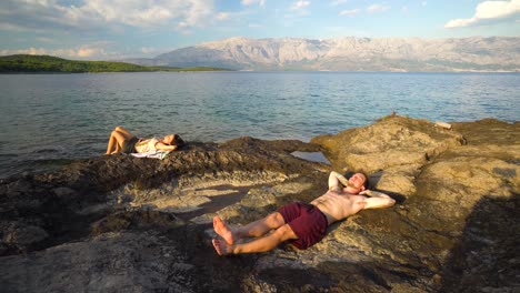 Un-Par-De-Turistas-Tomando-El-Sol-En-Las-Rocas-De-La-Playa-De-Sumartin-En-La-Isla-De-Brac,-Croacia,-Alrededor-De-Junio-De-2016.