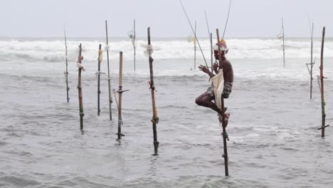 Old-man-alone-stilt-fishing-in-Weligama-beach-slow-motion-b-roll-clip,-also-rough-sea-waves-crashing-behind-him