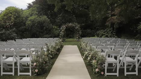 View-of-a-beautiful-flower-arch-placed-at-the-end-of-a-hallway-to-the-altar-in-a-venue-with-delicate-white-chairs-on-each-side-of-the-hallway