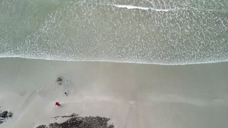 Luftaufnahmen-über-Einem-Fels--Und-Sandstrand-Mit-Vater-Und-Kind,-Die-Mit-Wellen-Spielen,-In-Der-Ferne-Surfer