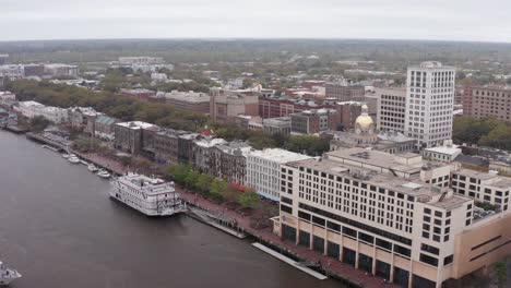 Descending-and-panning-aerial-shot-of-a-river-cruise-ship-docked-along-the-river-in-downtown-Savannah,-Georgia