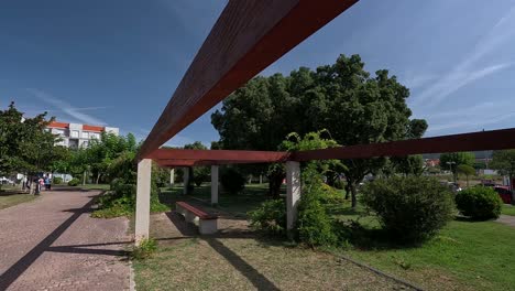 Estructura-De-Madera-Sin-Techo-Con-Soportes-De-Granito-En-Medio-Del-Parque-Público-Donde-Los-Turistas-Caminan-En-Verano-En-Un-Día-De-Sol-Y-Calor-Radiante,-Disparos-De-Grúas-Bajando,-Pontevedra,-Galicia,-España