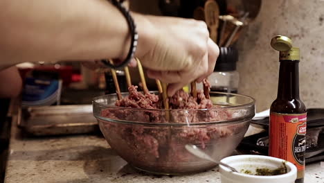 Caucasian-woman's-hands-mix-raw-ground-hamburger-meat-with-wooden-forks-in-glass-bowl,-side-close-up-view