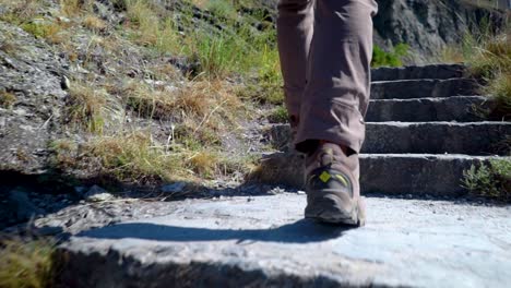 In-Zeitlupe-Folgt-Die-Untere-Hälfte-Eines-Mannes,-Der-An-Einem-Sonnigen-Tag-In-Wanderschuhen-Steinstufen-Hinaufgeht