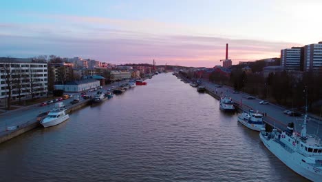 Aerial,-rising,-drone-shot-over-the-Aurajoki-river,-overlooking-boats-and-the-city-of-Turku,-during-sunset,-in-Varsinais-suomi,-Finland