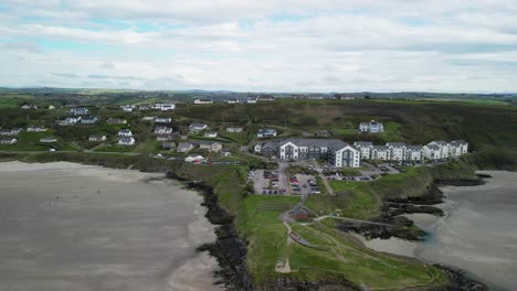 La-Playa-De-Clonakilty-Inchidoney-Con-El-Albergue-Y-El-Hotel-Spa,-El-Pueblo-Y-Las-Imágenes-Aéreas-Despegan-Hacia-Atrás,-Revelando-Una-Costa-Escarpada-Y-Una-Playa-De-Arena.