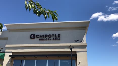 The-exterior-of-a-Chipotle-fast-casual-Tex-Mex-Restaurant-in-a-strip-mall-in-Portland,-Oregon,-USA-with-a-handheld-pan-down-through-some-trees