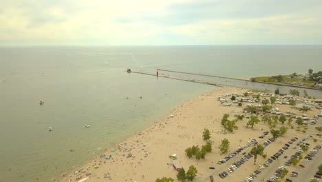 Luftaufnahme-Eines-Großen-Strandes-Mit-Einer-Menschenmenge-Neben-Einem-Campingplatz-Und-Parkplatz-Mit-Blick-Auf-Einen-Großen-See-Mit-Einem-Pier-Und-Einem-Leuchtturm