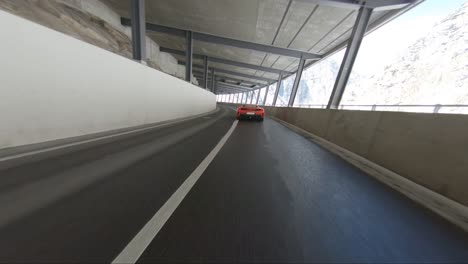 Ferrari-in-the-alps-driving-fast-along-a-mountain-pass-tunnel-viewed-with-a-drone-shot-from-outside-the-tunnel-to-right-next-to-the-car