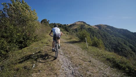 Ciclista-De-Montaña-Masculino-En-Bicicleta-Cuesta-Arriba-En-Camino-De-Grava,-Mirando-Hacia-Las-Montañas-En-Un-Día-Soleado-De-Verano,-Tiro-Amplio-En-Cámara-Lenta