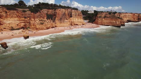 Coastline-view-with-beachgoers-and-beautiful-orange-rugged-cliffs-by-aerial-4k-drone-at-Estrada-da-Caramujeira-in-the-Algarve-region-of-Portugal