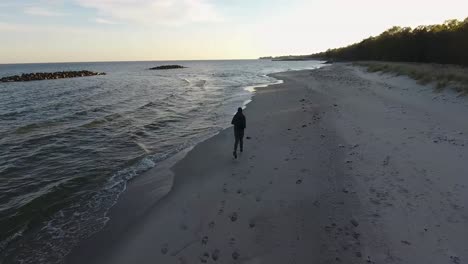 Aerial-Tracking-Shot-of-a-Man-Running-By-the-Östersjö-Ocean-Waves-at-Ystad-Saltsjöbad-Beach-in-South-Sweden-Skåne-in-The-Summer-Evening
