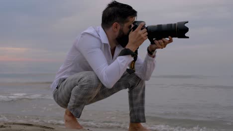 Ein-Fotograf,-Der-Bilder-Macht,-Während-Er-Am-Strand-Hockt-Und-Seine-Füße-Im-Wasser-Hält