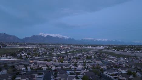 Twilight-over-a-suburban-sprawl-in-a-valley-beneath-the-snow-capped-mountains---pullback-aerial-reveal