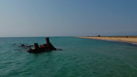 Paning-revealing-aerial-drone-shot-of-a-shipwreck-that-extends-into-the-ocean-over-a-golden-sandy-shore-and-the-blue-sea-of-Mediterranean-sea-close-to-Thessaloniki,-Greece-in-Epanomi