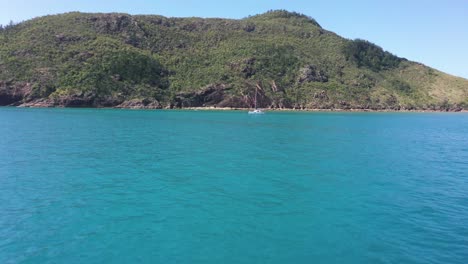 Antena-Baja-Sobre-El-Mar-Azul-Hasta-El-Velero-Catamarán-Blanco-Amarrado-Frente-A-La-Isla