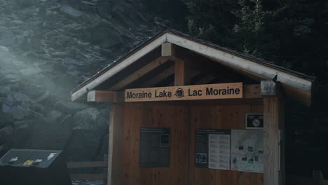 Pequeña-Estructura-De-Madera-Con-Señalización-Del-Lago-Morrena-En-El-Parque-Nacional-Banff-En-Alberta,-Canadá,-Revelada-Detrás-Del-Efecto-Bokeh-En-Un-Día-Soleado-Y-Brillante---Toma-Panorámica