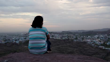 Isoliertes-Mädchen,-Das-Die-Stadtlandschaft-Auf-Einem-Berggipfel-Mit-Dramatischem-Himmel-In-Der-Abenddämmerung-Beobachtet.-Das-Video-Wurde-In-Mehrangarh,-Jodhpur,-Rajasthan,-Indien-Aufgenommen