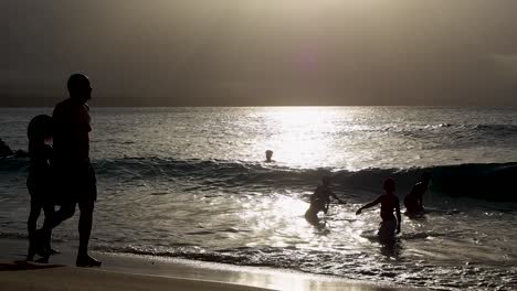 Strand-Bei-Sonnenuntergang-Mit-Silhouetten-Von-Menschen,-Die-Im-Wasser-Spielen